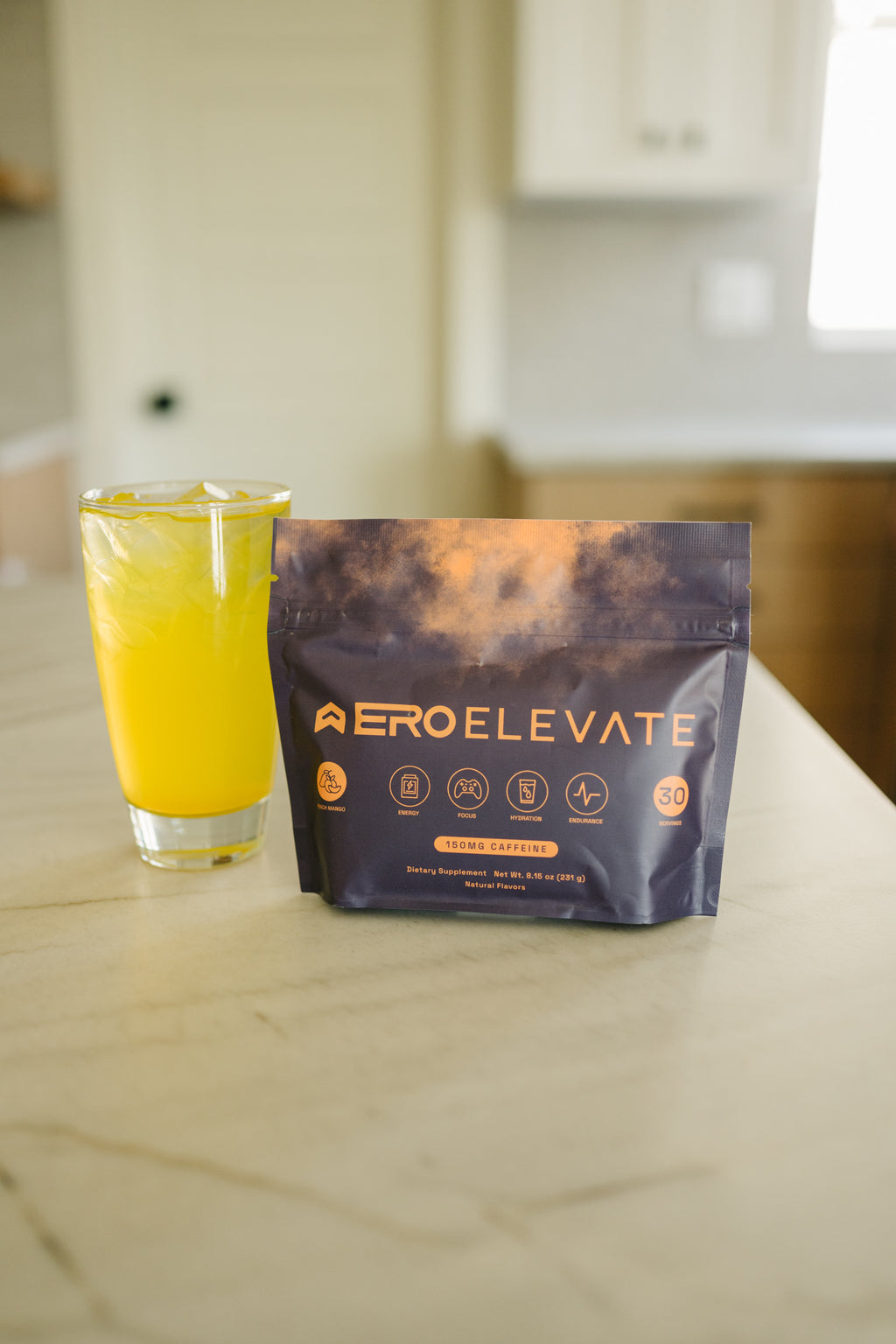 ERO ELEVATE Peach Mango Caffeine Drink Mix - Dietary Supplement for Energy, Focus, Hydration, and Endurance. Natural flavors with 150mg caffeine per serving, shown in vibrant orange drink next to resealable pouch on a kitchen counter.